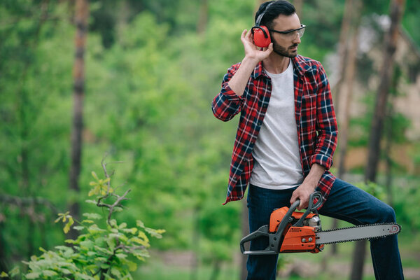 lumberjack in plaid shirt standing with chainsaw in forest and touching noise-canceling headphones