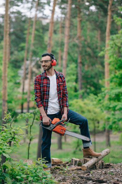 smiling lumberman in plaid shirt and denim jeans standing with chainsaw in forest and looking away