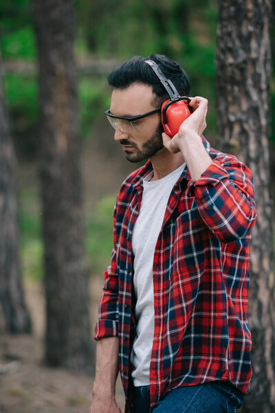 задумчивый лесоруб в защитных очках, стоящих в лесу и трогательных шумоподавляющих наушниках
