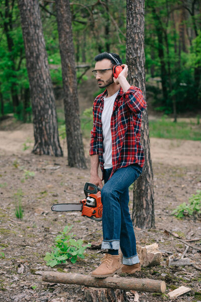 thoughtful lumberjack holding chainsaw, touching noise-canceling headphones and looking away