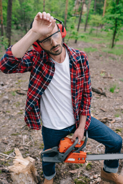 exhausted lumberjack with chainsaw holding hand near head and looking away in forest