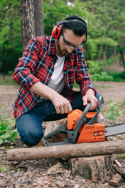 lumberman in plaid shirt and hearing protectors adjusting chainsaw in forest