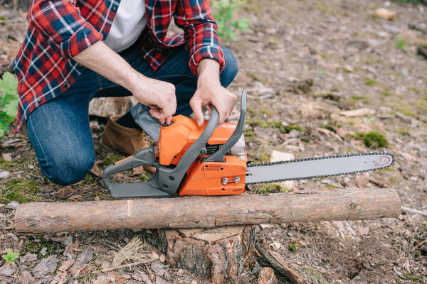 partial view of lumberjack adjusting chainsaw while sitting near tree trunk in forest