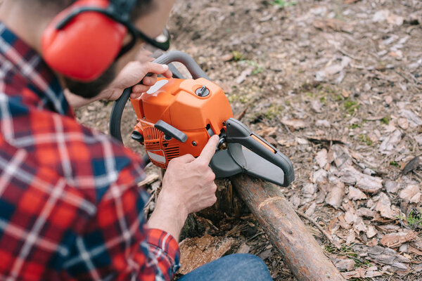 selective focus of lumberjack in hearing protectors repairing chainsaw in forest