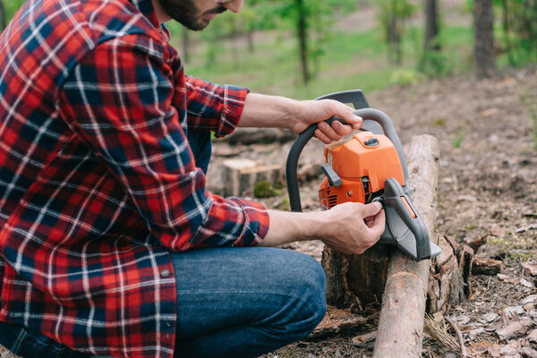 cropped view of lumberjack in plaid shirt repairing chainsaw in forest