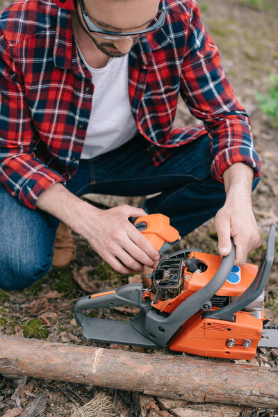 partial view of lumberman in plaid shirt adjusting chainsaw in forest