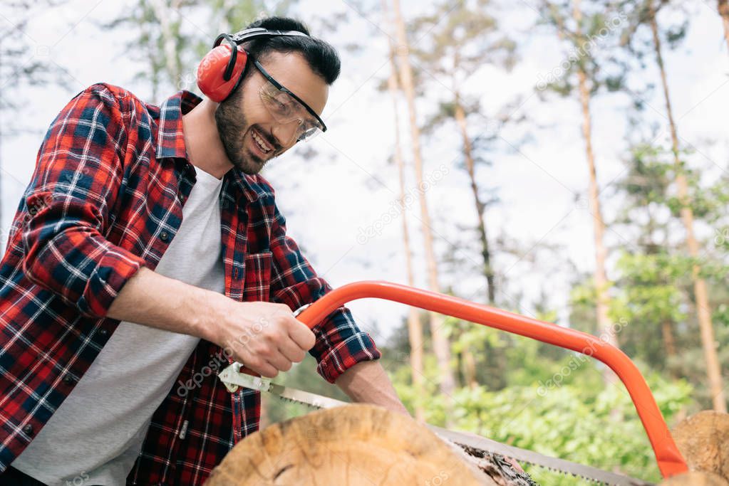 smiling lumberer in earmuffs and protective glasses cutting log with handsaw in forest