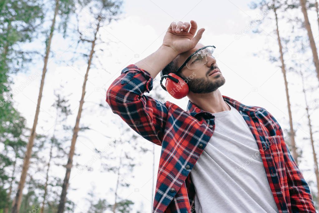 tired lumberjack in protective glasses holding hand near head and looking away in forest