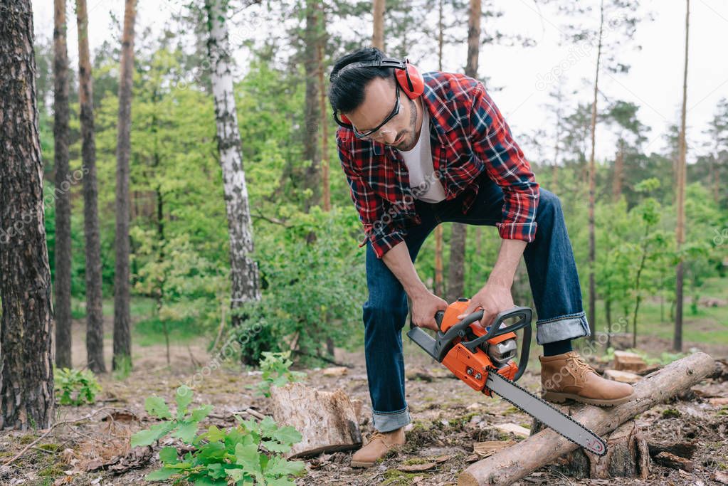 lumberjack in plaid shirt and denim jeans cutting log with chainsaw in forest