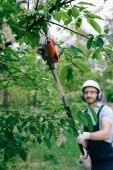 selektivní zaměření usmívajícího se zahradníka v helmě a chrániče sluchu zastřihování stromů s teleskopickou pilou a s pohledem na fotoaparát