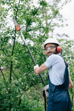 smiling gardener in helmet trimming trees with telescopic pole saw and looking at camera clipart