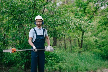 cheerful gardener in overalls, helmet and hearing protectors holding telescopic pole saw and smiling at camera clipart