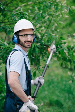 cheerful gardener in helmet, protective glasses and noise-canceling headphones holding telescopic pole saw and smiling at camera clipart