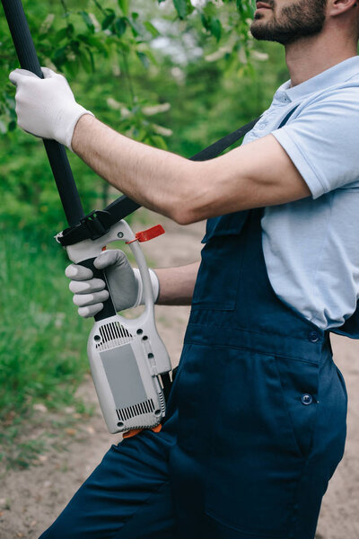 partial view of gardener in overalls trimming trees with telescopic pole saw in garden