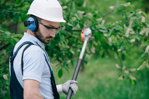 selective focus of pensive gardener in helmet and hearing protectors trimming trees with telescopic pole saw in park