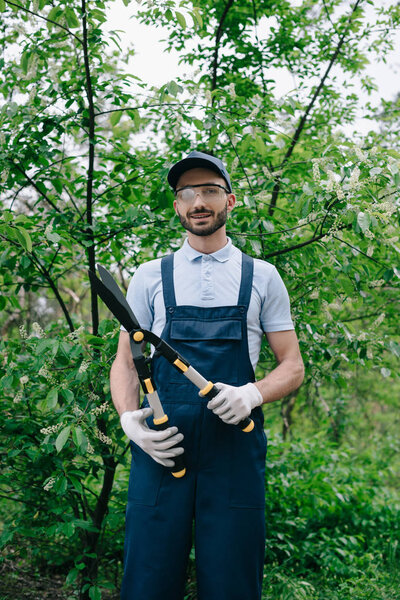 handsome gardener in overalls, cap and protective glasses holding trimmer and smiling at camera