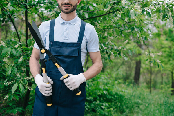 partial view of smiling gardener in overalls holding trimmer in garden