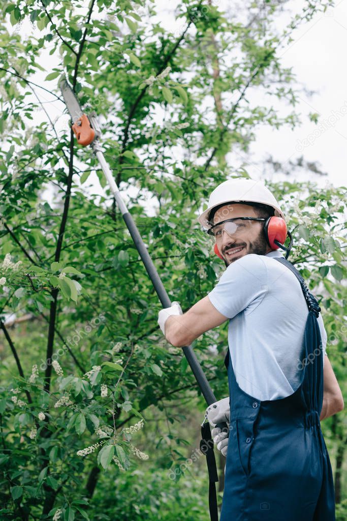 smiling gardener in helmet trimming trees with telescopic pole saw and looking at camera