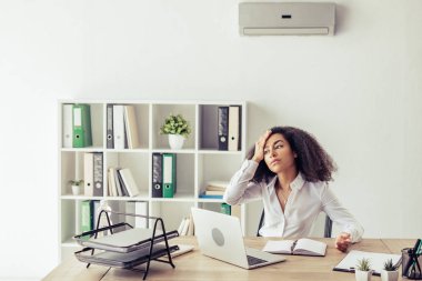 atttractive african american businesswoman sitting at workplace under air conditioner and suffering from heat clipart