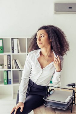 exhausted african american businesswoman sitting on desk while suffering from heat in office clipart