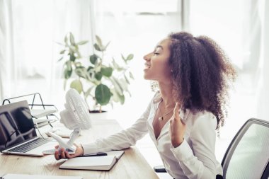 ofiste işyerinde otururken elektrik fan üfleme tutarak neşeli afrikalı-amerikalı iş kadını