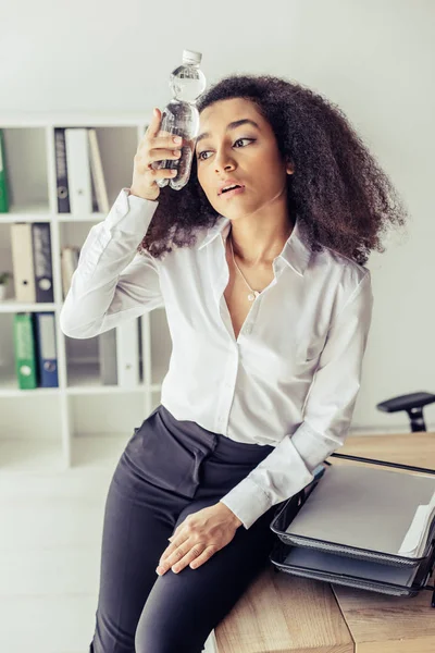 精疲力竭的非洲裔美国女商人拿着塑料瓶 靠近头部 而在办公室遭受热 — 图库照片