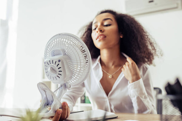 selective focus of attractive african american businesswoman holding electric fan while sitting at workplace 