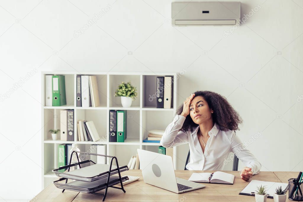 atttractive african american businesswoman sitting at workplace under air conditioner and suffering from heat