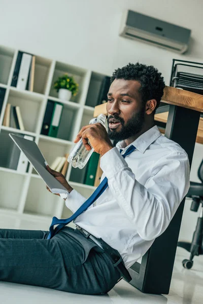 若いアフリカ系アメリカ人のビジネスマンがオフィスで暑さに苦しみながら 床に座って水を飲む — ストック写真