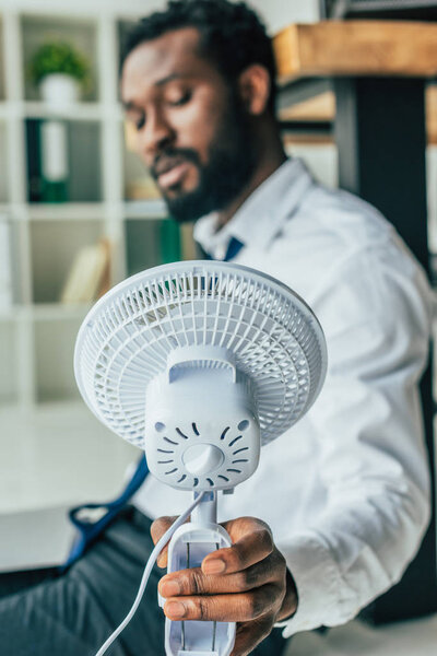 selective focus of african american businessman sitting on floor and holding electric fan