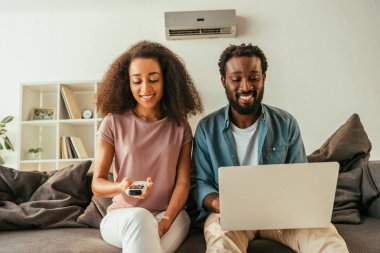 dizüstü bilgisayar kullanarak neşeli afro-amerikan adam yakınında otururken uzaktan kumanda tutan gülümseyen afrikalı amerikalı kadın