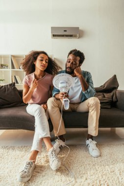 genç afrikalı amerikalı erkek ve kadın kanepede oturan ve evde ısı muzdarip iken elektrik fan tutarak