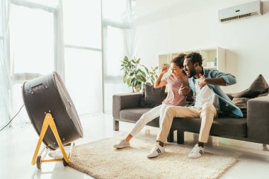 heyecanlı afro-amerikan erkek ve kadın elektrikli fan üfleme önünde kanepede oturan