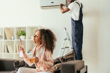 düşünceli afro-amerikan kadın dergi ve çay fincan ile kanepede otururken, afro-amerikan handyman klima tamir ederken