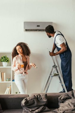 mutsuz afro-amerikan kadın uzaktan kumanda ve çay fincan tutan handyman yanında dururken klima tamir