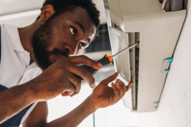 serious african american repairman fixing air conditioner with screwdriver clipart