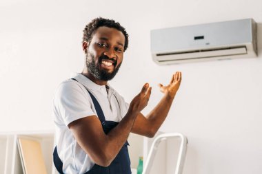 cheerful african american repairman smiling at camera and pointing with hands at air conditioner clipart