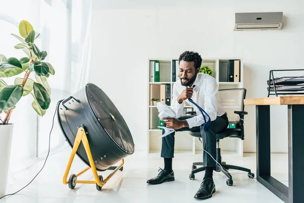 Афроамериканець Бізнесмен Тримає Стіл Вентилятор Сидячи Перед Електричним Вентилятором Офісі — стокове фото