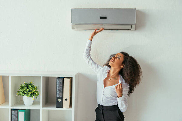 довольно африканская американская бизнесвумен трогает кондиционер, страдая от жары в офисе
