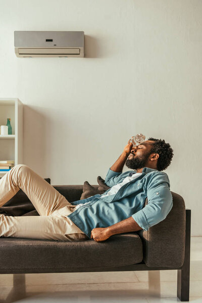 african american man lying on sofa with closed eyes and holding bottle with water near head