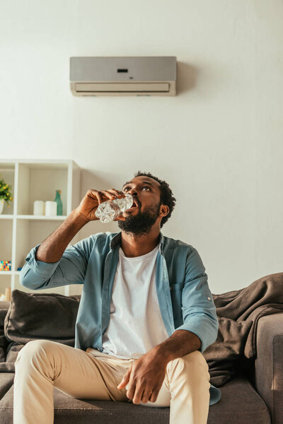thirsty african american man drinking water from plastic bottle while sitting on sofa at home