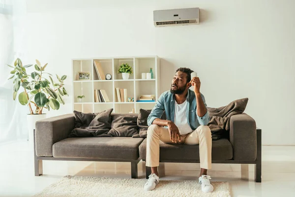 Unavený Afroamerický Muž Sedící Pohovce Přidržuje Dálkový Ovladač Klimatizace Zatímco — Stock fotografie