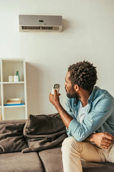 Africano Americano Homem Segurando Condicionado Controlador Remoto Enquanto Sentado Sofá — Fotografia de Stock