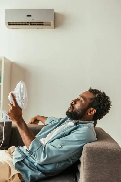 Hombre Afroamericano Sosteniendo Soplado Ventilador Eléctrico Mientras Está Acostado Sofá —  Fotos de Stock