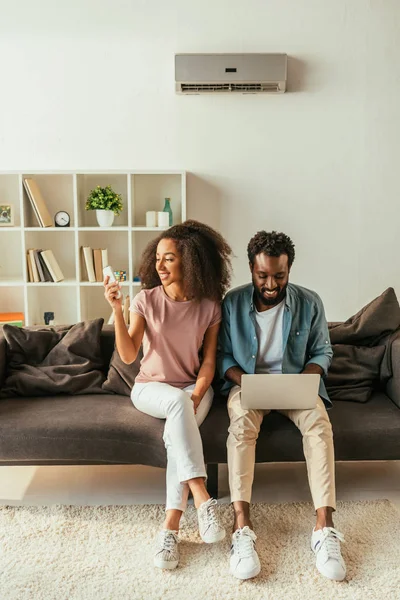 Glimlachende Afrikaanse Vrouw Met Air Conditioner Afstandsbediening Terwijl Zittend Bank — Stockfoto