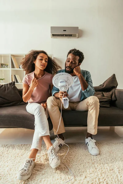 Hombre Mujer Afroamericanos Jóvenes Sentados Sofá Sosteniendo Ventilador Eléctrico Mientras — Foto de Stock