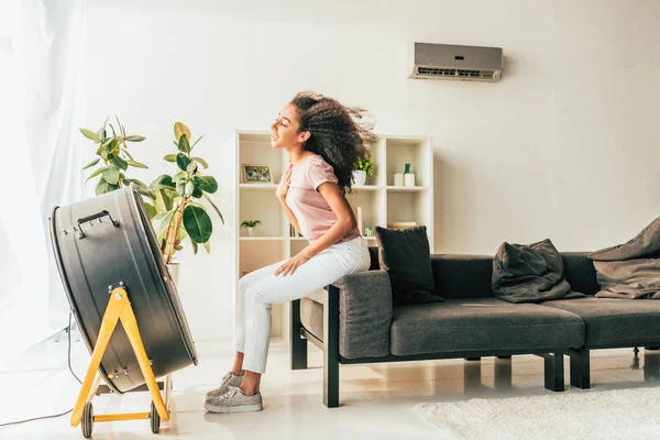 Sorrindo Afro Americano Mulher Sentada Frente Ventilador Sopro Elétrico Casa — Fotografia de Stock