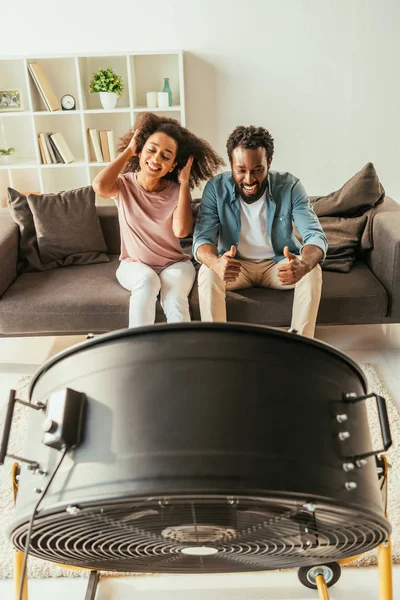 happy african american man showing thumbs up while sitting near cheerful african american woman in front if blowing electric fan