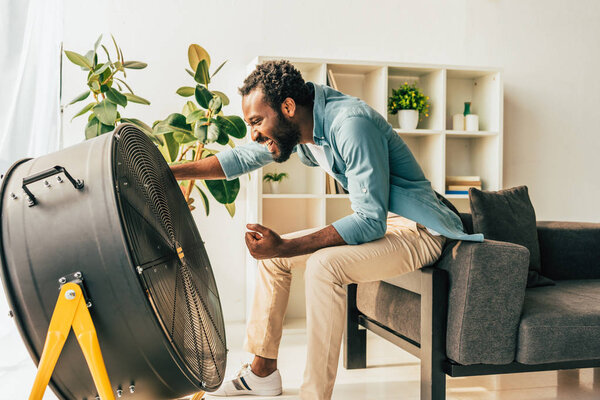 excited african american man sitting near electric ventilator at home