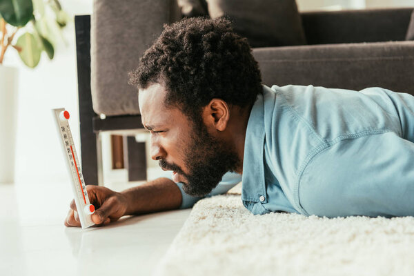 shocked african american man looking at thermometer while lying on floor and suffering from summer heat